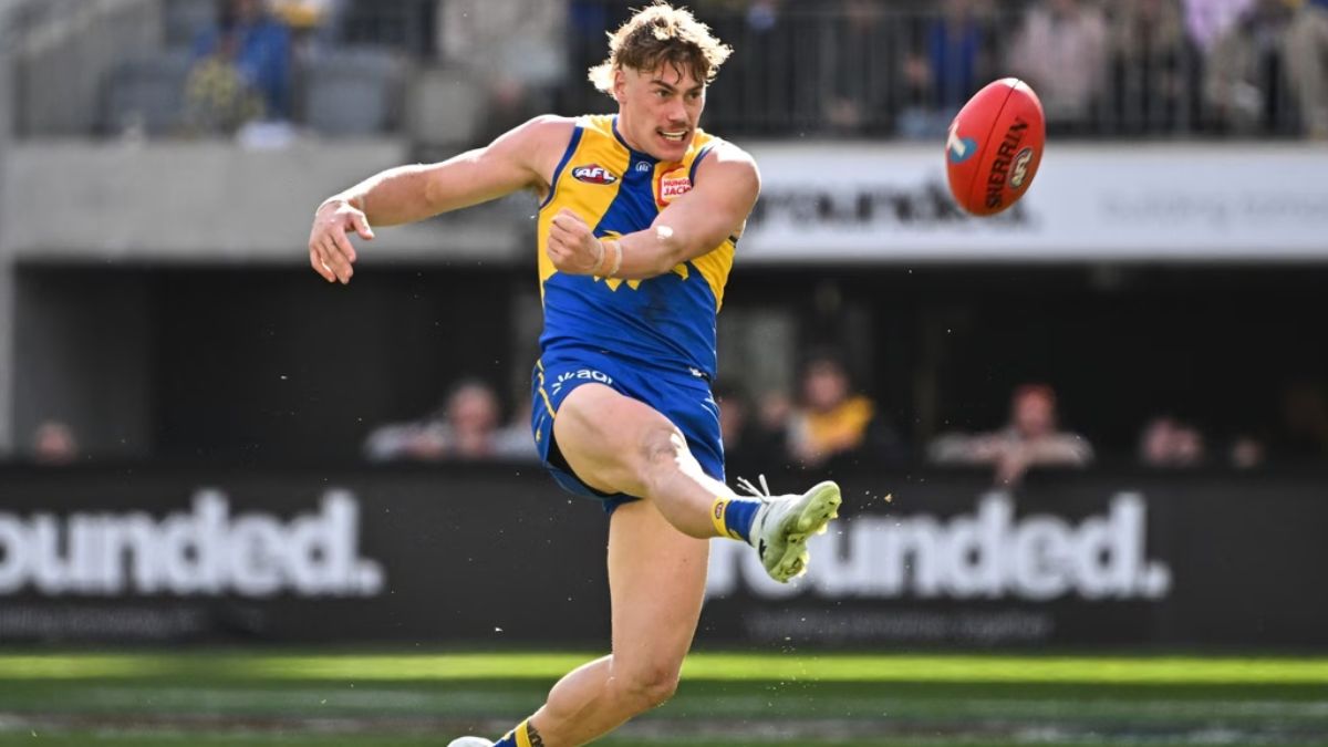 Male Australian rules footballer mid-kick in blue-and-yellow uniform, ball flying ahead on a sunny field.