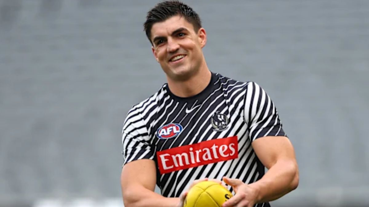 Male athlete in a black-and-white striped AFL jersey holding a yellow football, smiling at the camera with a gray stadium background.