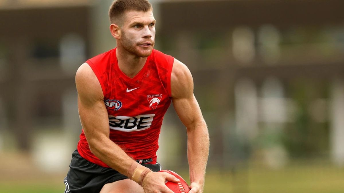 Male Australian rules football player in a red sleeveless training jersey, holding a football during practice on a field.