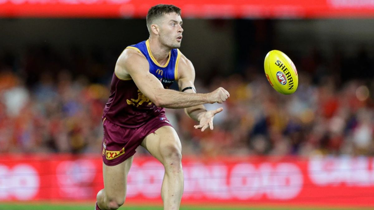 Australian rules footballer in maroon and gold uniform reaches toward a yellow Sherrin ball during a match with a blurred crowd in the background.