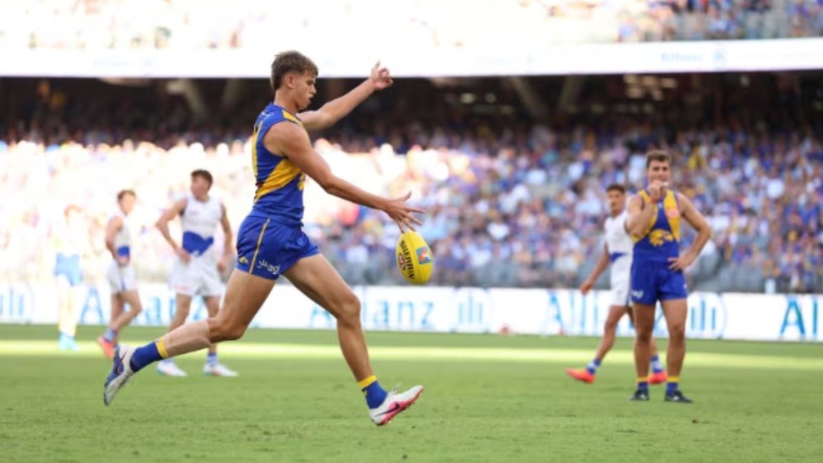 Australian rules footballer in blue and yellow jersey kicks a ball on a green field with a stands-full crowd in the background.