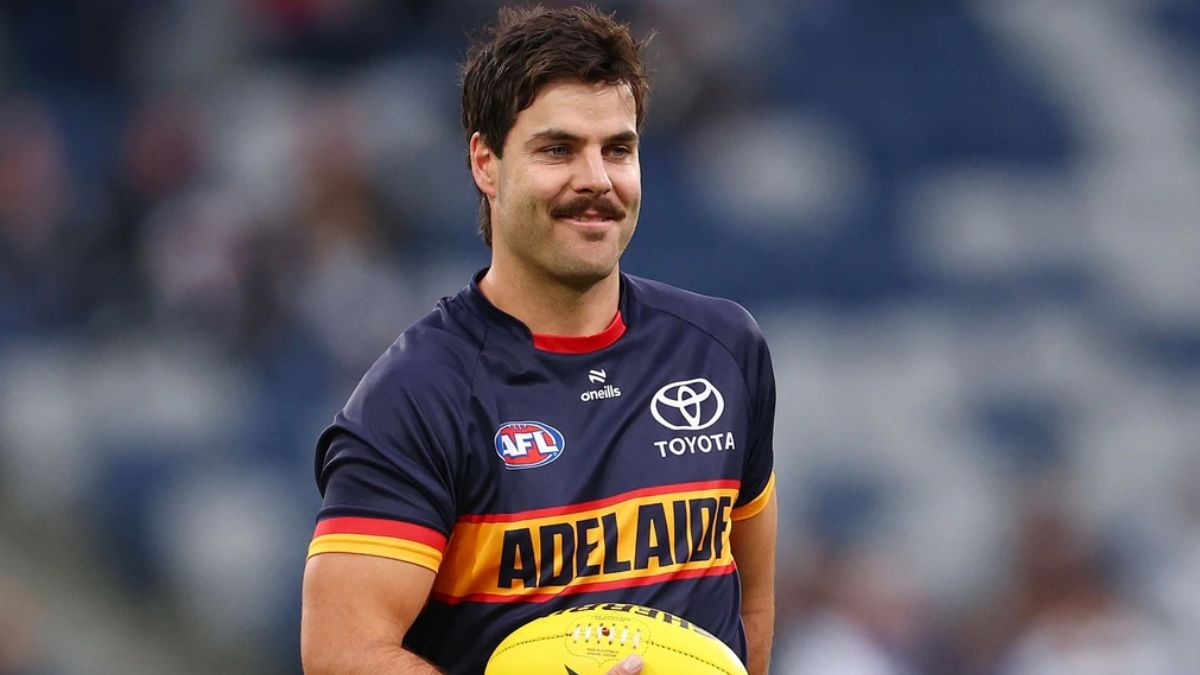 A male AFL player wearing an Adelaide Crows navy jersey with red-yellow trim, holding a yellow football on the field.